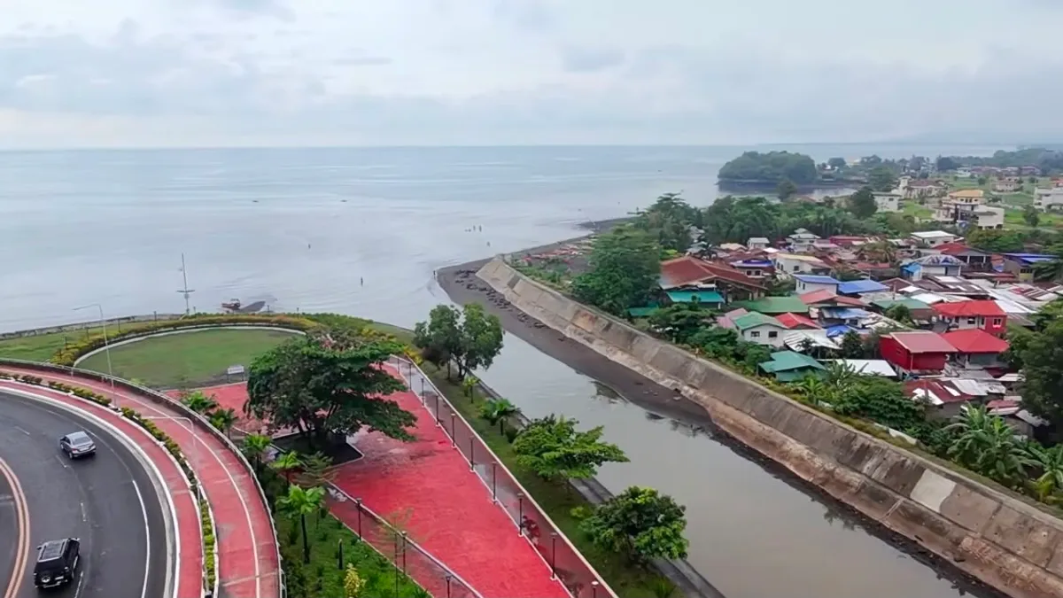 Aerial view of the Davao Coastal Road curving past a river mouth, with a dense low-rise barangay along the far bank